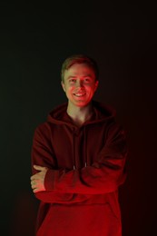 Photo of Portrait of smiling man wearing hoodie with crossed arms in neon lights against black background