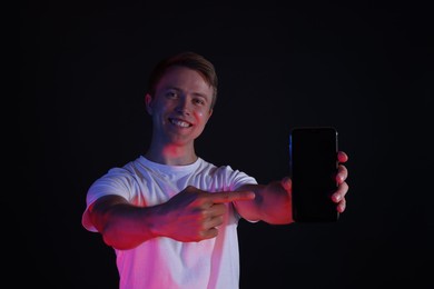 Photo of Smiling man pointing at smartphone in neon lights against black background