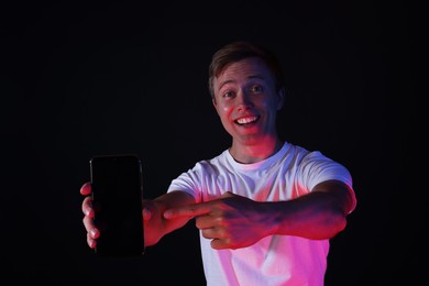 Photo of Smiling man pointing at smartphone in neon lights against black background