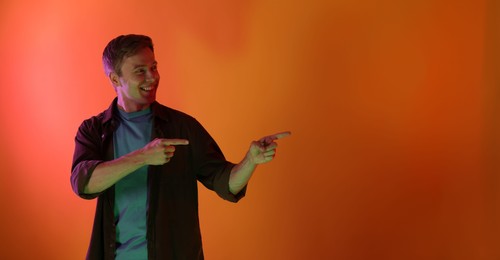 Photo of Smiling man pointing at something in neon lights against orange background. Space for text