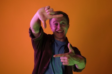 Photo of Smiling man showing frame gesture in neon lights against orange background