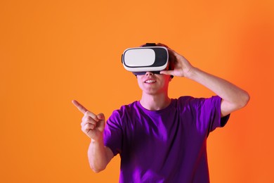 Photo of Smiling man using virtual reality headset and pointing at something in neon lights against orange background. Space for text