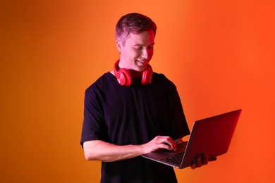 Photo of Smiling man with headphones using laptop in neon lights against orange background