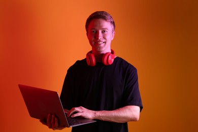 Photo of Portrait of smiling man with headphones using laptop in neon lights against orange background
