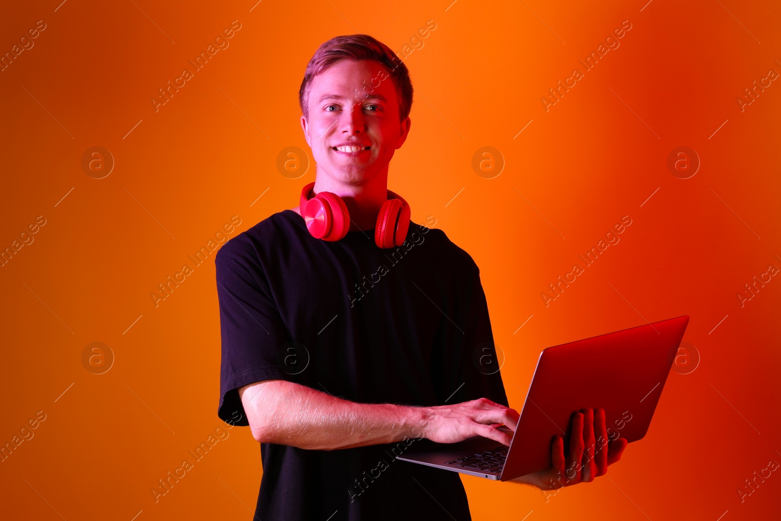 Portrait of smiling man with headphones using laptop in neon lights against orange background Photo of Portrait of smiling man with headphones using laptop in neon lights against orange background