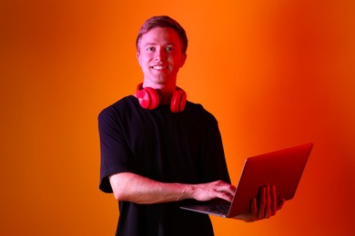 Photo of Portrait of smiling man with headphones using laptop in neon lights against orange background