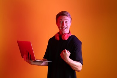 Photo of Happy man with headphones using laptop in neon lights against orange background