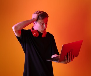 Photo of Emotional man with headphones using laptop in neon lights against orange background