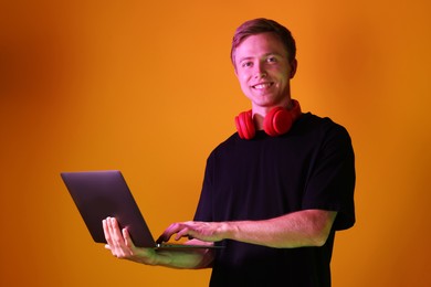 Photo of Portrait of smiling man with headphones using laptop in neon lights against orange background
