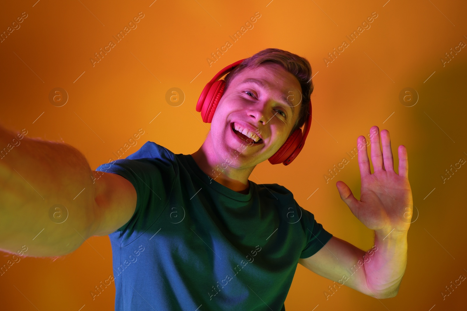 Smiling man wearing headphones taking selfie and waving hello in neon lights against orange background Photo of Smiling man wearing headphones taking selfie and waving hello in neon lights against orange background