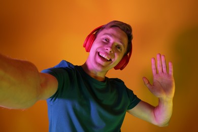 Photo of Smiling man wearing headphones taking selfie and waving hello in neon lights against orange background