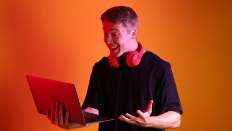Photo of Happy man with headphones using laptop in neon lights against orange background