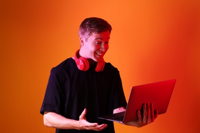 Photo of Happy man with headphones using laptop in neon lights against orange background