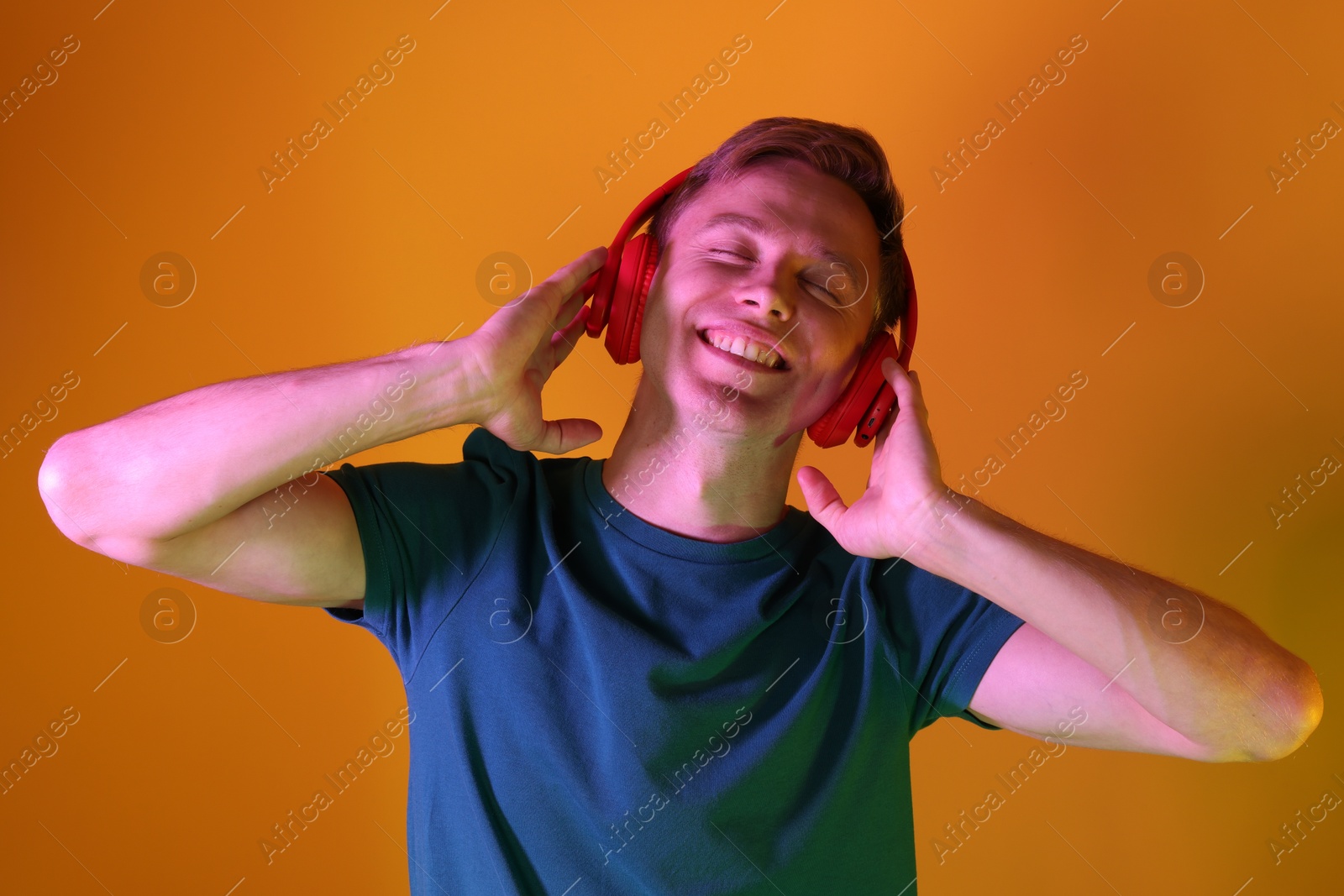 Smiling man wearing headphones listening to music in neon lights against orange background Photo of Smiling man wearing headphones listening to music in neon lights against orange background