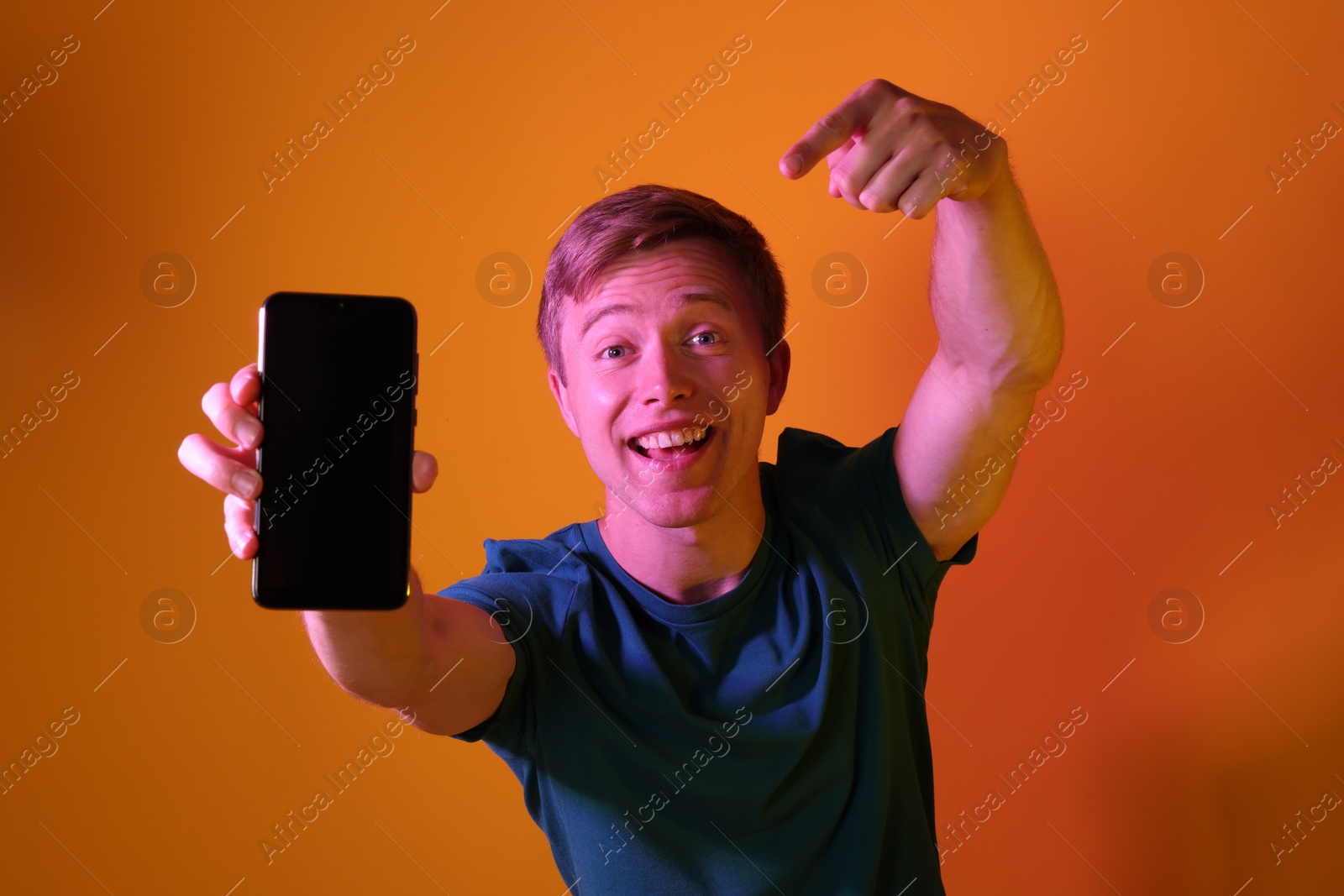 Photo of Happy man pointing at smartphone in neon lights against orange background