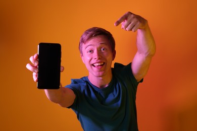 Photo of Happy man pointing at smartphone in neon lights against orange background
