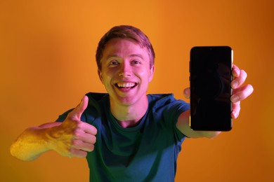 Photo of Happy man with smartphone showing thumbs up in neon lights against orange background