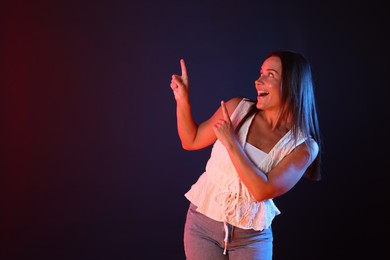 Photo of Smiling woman in neon lights pointing upwards against dark background. Space for text