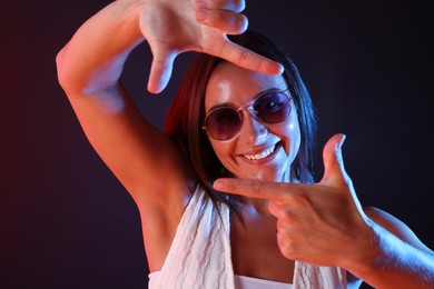 Photo of Happy woman wearing sunglasses showing frame gesture in neon lights against dark background