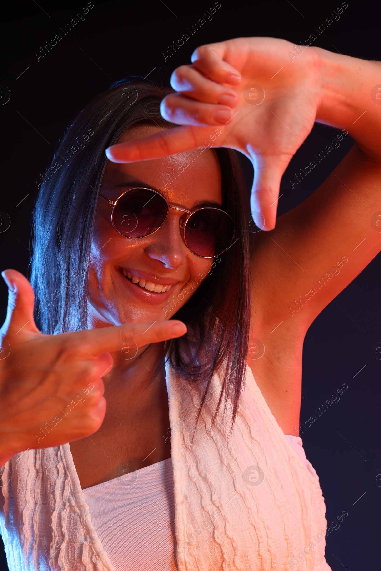 Happy woman wearing sunglasses showing frame gesture in neon lights against dark background Photo of Happy woman wearing sunglasses showing frame gesture in neon lights against dark background