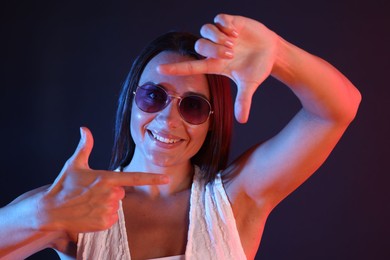 Photo of Happy woman wearing sunglasses showing frame gesture in neon lights against dark background