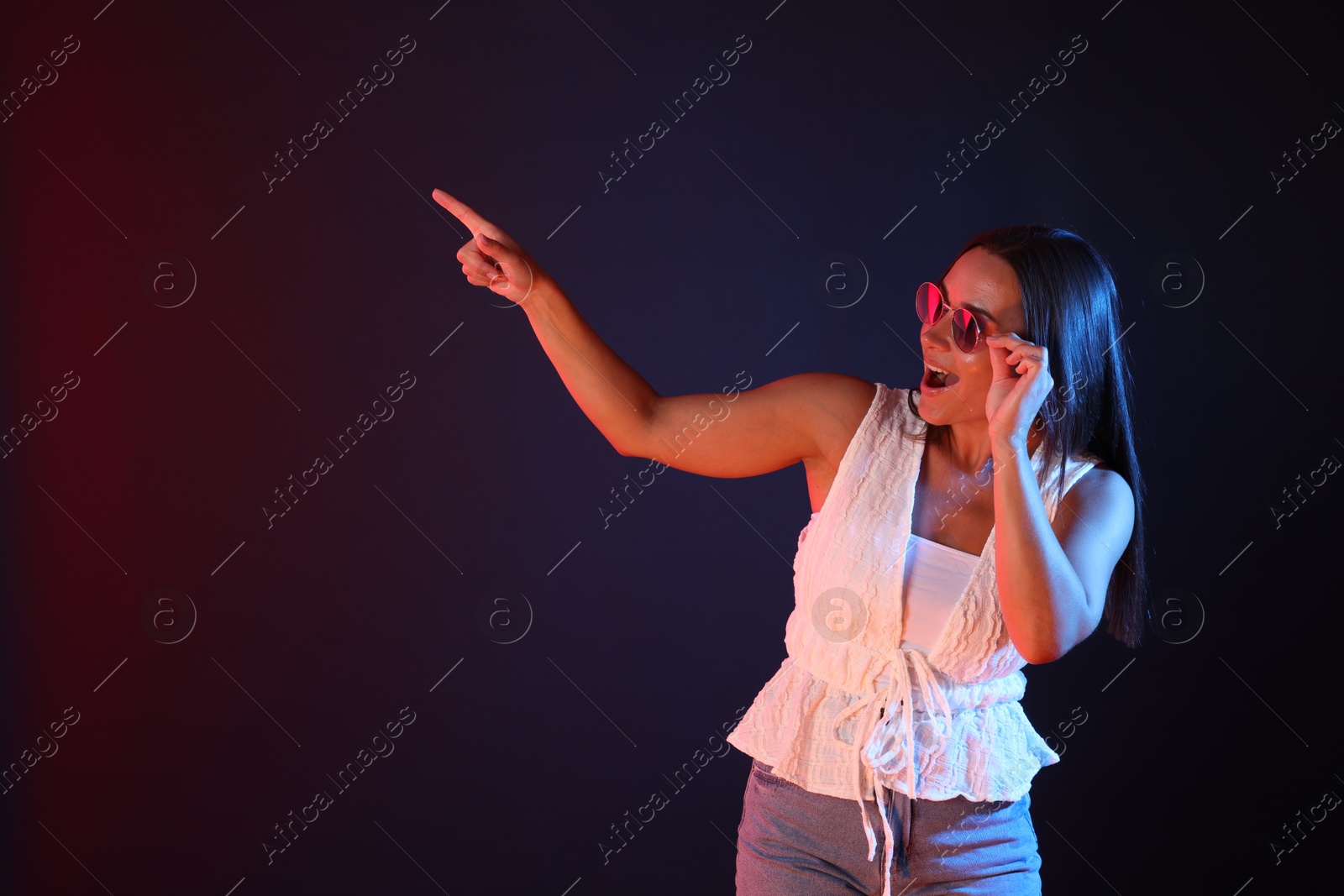 Excited woman wearing sunglasses in neon lights pointing at something against dark background. Space for text Photo of Excited woman wearing sunglasses in neon lights pointing at something against dark background. Space for text