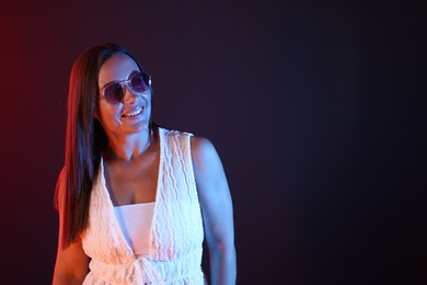 Photo of Happy woman wearing sunglasses in neon lights against dark background. Space for text