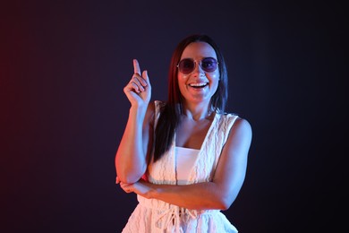 Photo of Smiling woman wearing sunglasses pointing upwards in neon lights against dark background