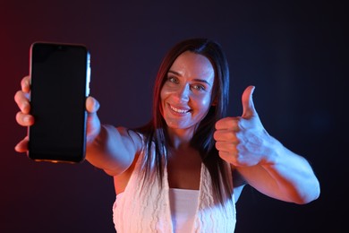 Photo of Happy woman showing smartphone and thumbs up in neon lights against dark background