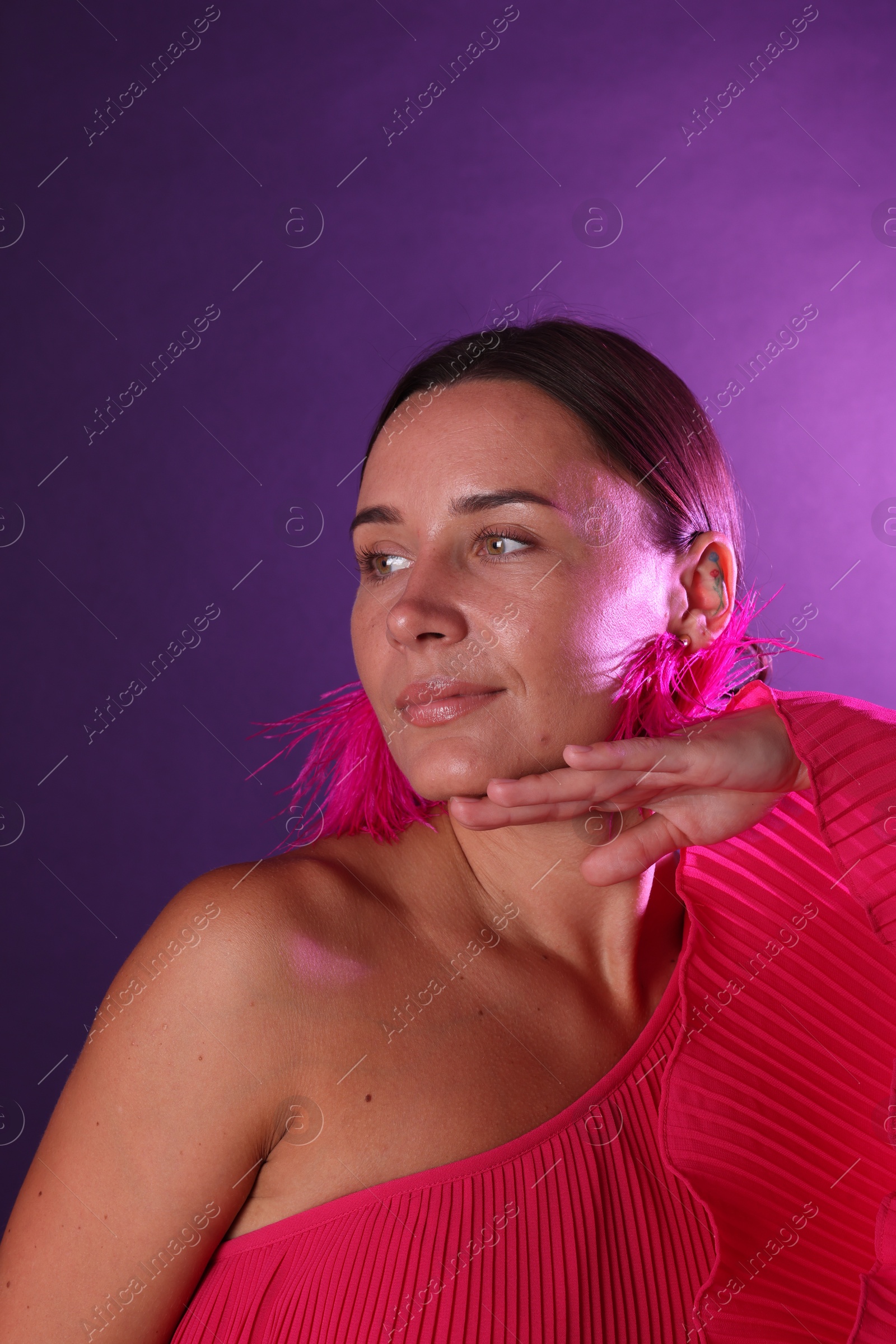 Beautiful woman wearing fur earrings in neon lights against purple background Photo of Beautiful woman wearing fur earrings in neon lights against purple background