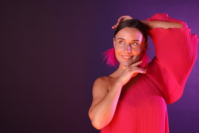 Photo of Smiling woman wearing fur earrings in neon lights against purple background. Space for text