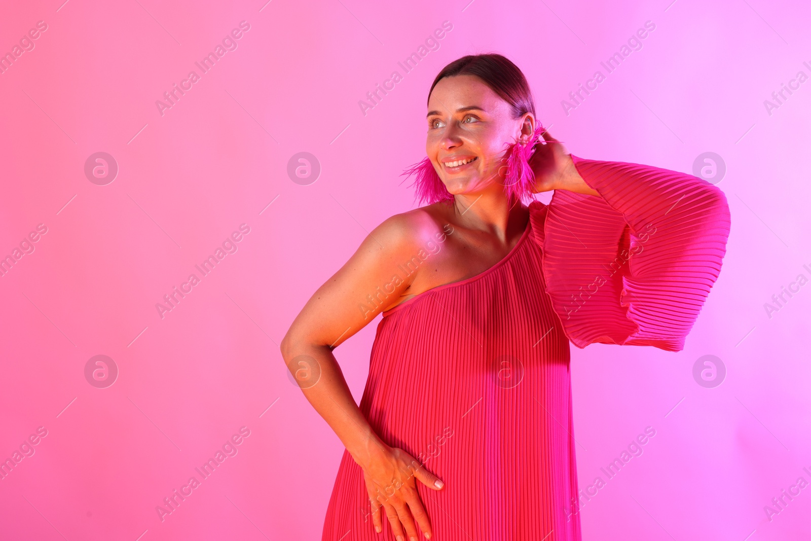 Smiling woman wearing fur earrings in neon lights against pink background Photo of Smiling woman wearing fur earrings in neon lights against pink background
