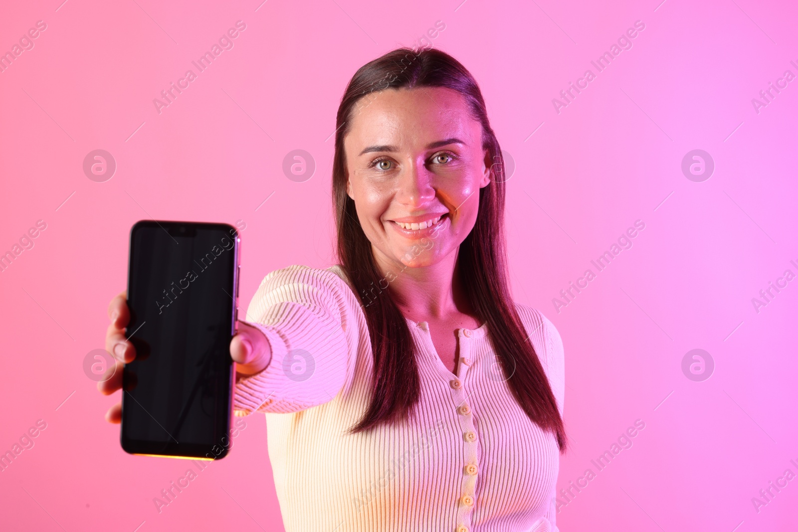 Photo of Smiling woman showing smartphone in neon lights against pink background