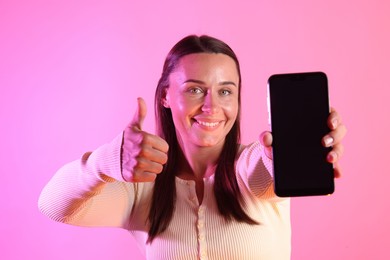 Photo of Smiling woman showing smartphone and thumbs up in neon lights against pink background