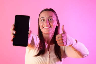 Photo of Smiling woman showing smartphone and thumbs up in neon lights against pink background