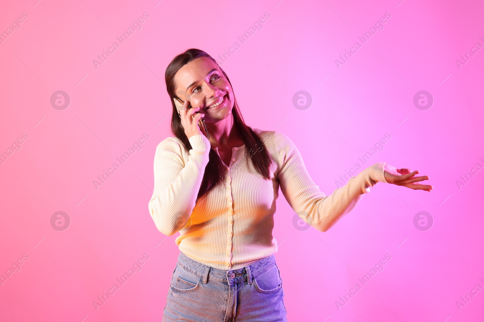 Photo of Smiling woman talking by smartphone in neon lights against pink background