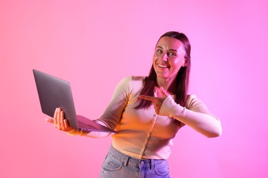 Photo of Smiling woman pointing at laptop in neon lights against pink background