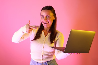 Photo of Smiling woman pointing at laptop in neon lights against pink background