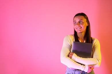 Photo of Smiling woman with laptop in neon lights against pink background. Space for text