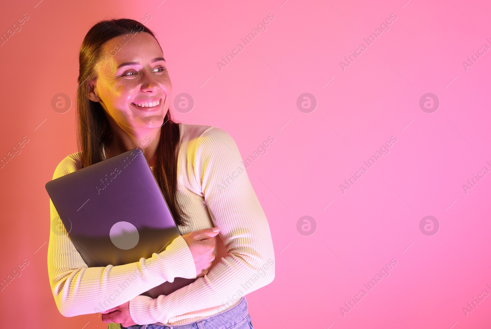 Smiling woman with laptop in neon lights against pink background. Space for text Photo of Smiling woman with laptop in neon lights against pink background. Space for text