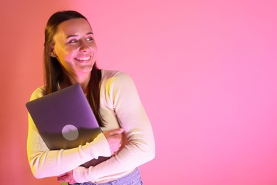 Photo of Smiling woman with laptop in neon lights against pink background. Space for text