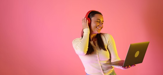 Photo of Smiling woman in headphones with laptop in neon lights against pink background. Space for text