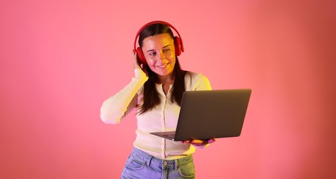 Photo of Smiling woman in headphones with laptop in neon lights against pink background