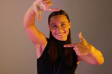 Photo of Smiling woman showing frame gesture in neon lights against grey background