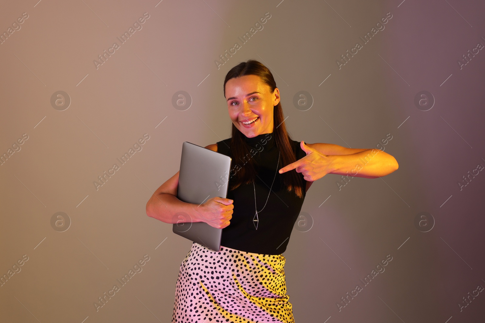 Happy woman pointing at laptop in neon lights against grey background Photo of Happy woman pointing at laptop in neon lights against grey background