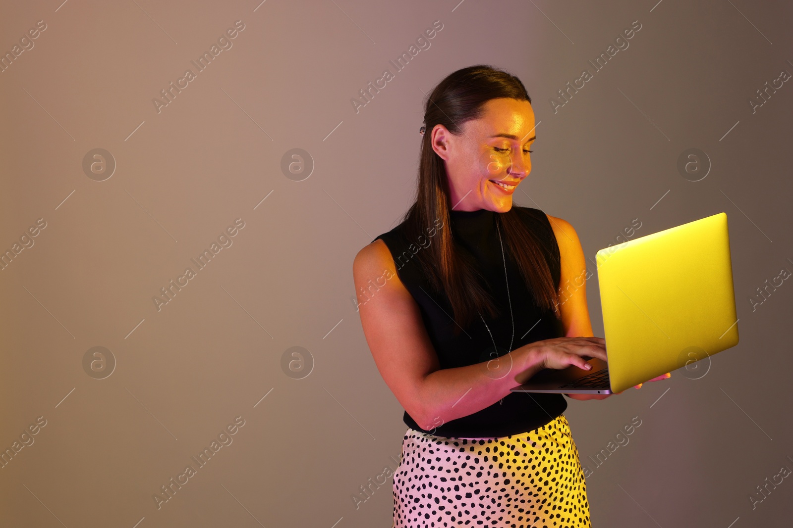 Photo of Smiling woman using laptop in neon lights against grey background. Space for text