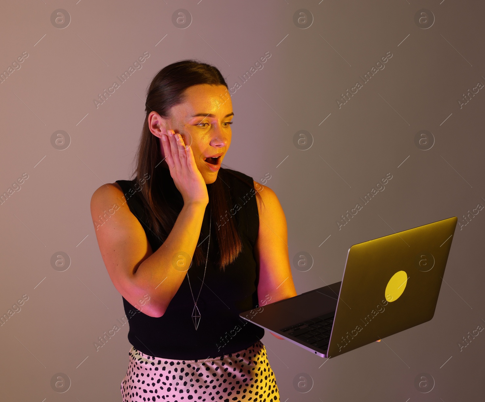 Emotional woman with laptop in neon lights against grey background Photo of Emotional woman with laptop in neon lights against grey background