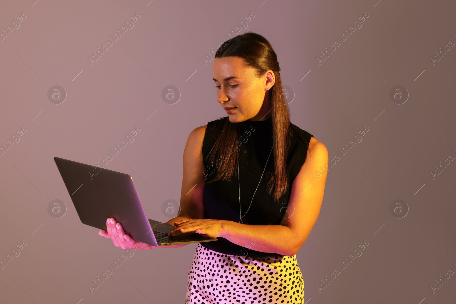Beautiful woman using laptop in neon lights against grey background Photo of Beautiful woman using laptop in neon lights against grey background