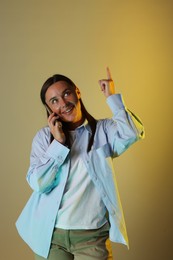 Photo of Smiling woman talking by smartphone and pointing upwards in neon lights against color background