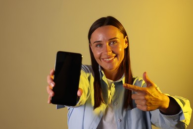 Photo of Smiling woman pointing at smartphone in neon lights against color background